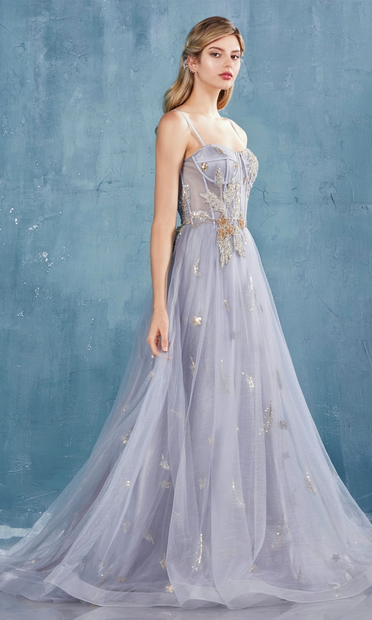 A woman stands in profile wearing the Shoulder-Bow Long Corset Prom Ball Gown A0824 with gold embellishments and thin straps, set against a textured blue background.